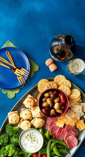 An arranged charcuterie board of veggies, meats, crackers and dips sits on a blue counter that has plates, a champagne bottles and glass flutes.