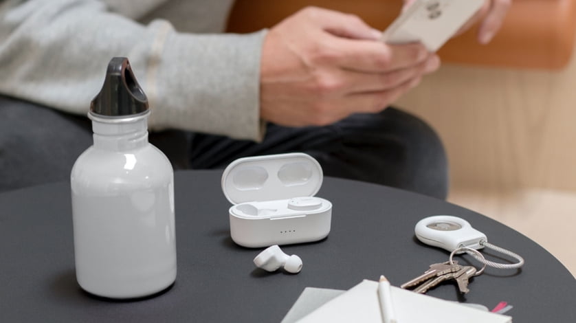 Wireless ear buds in the foreground on a table with a blurred out hand holding a cell phone in the background.