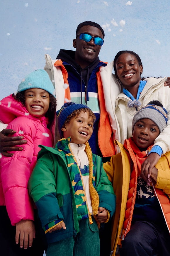 A smiling family of two adults and three children wear colorful winter jackets, hats and layers, standing close together.