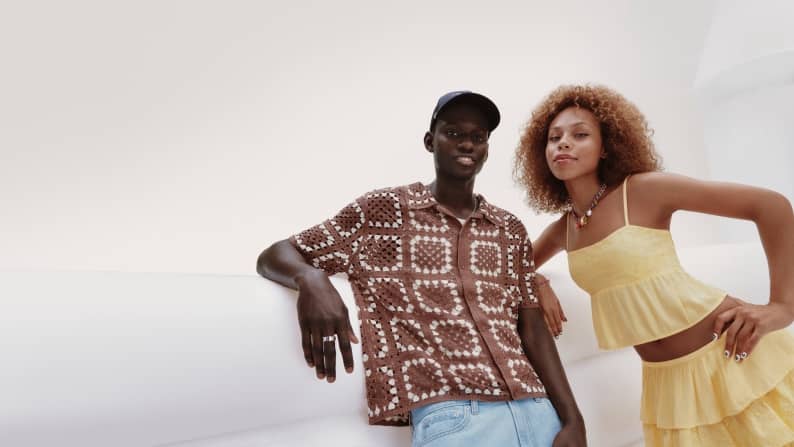 Young man wearing a brown patterned short sleeve shirt and cap standing with a young woman in a yellow crop top and matching skirt with necklace.