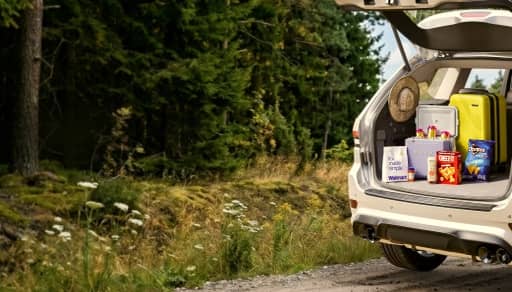 An open hatchback car filled with snacks, meds, skincare, accessories, and a suitcase is in a woodsy area.