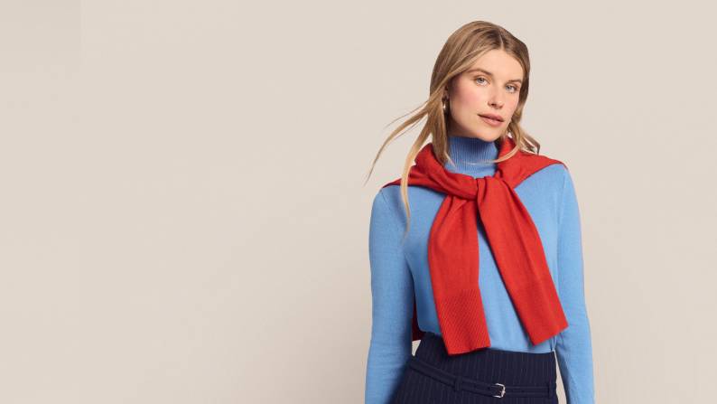 A woman wearing a blue sweater with a red sweater tied around her shoulder is pictured on a beige backdrop.