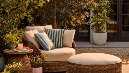 Cozy outdoor patio with wicker chair and ottoman, striped pillows, side table with drink, potted plants, and sunlit trees.