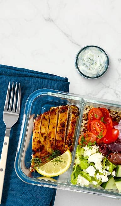 Grilled chicken, brown rice and fresh veggies are displayed in a glass storage container. A cup of yogurt dipping sauce is also shown.