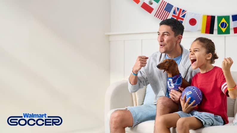 Walmart Soccer. Two people and a dog are cheering on a soccer game. They are dressed for the World Cup with a multi-country flag banner behind them.