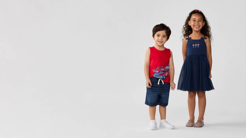 A little boy wearing a tank top and shorts and a little girl wearing a blue dress are standing together.