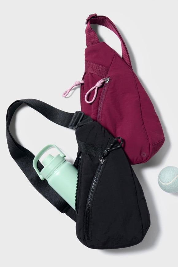 Black sling gym bag holding a mint green water bottle beside a magenta sling bag and tennis ball on a grey backdrop.