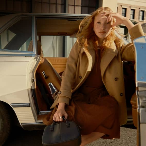 A woman is shown wearing a trench coat jacket over a dress. She is holding onto a small black handbag.