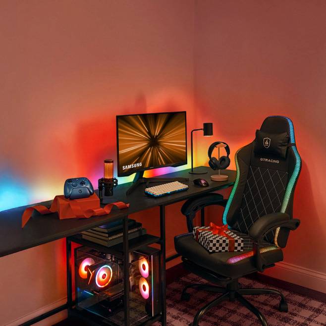 Samsung monitor and GT Racing gaming chair at RGB lit desk with keyboard, mouse, headset, controller, camera and gift boxes.