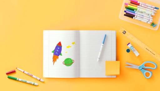 An open notebook with colorful drawings on white paper, next to markers, scissors and glue.