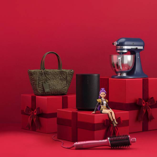 Green quilted handbag, black speaker, fashion doll and pink heated hair brush arranged with blue stand mixer on red gift boxes.
