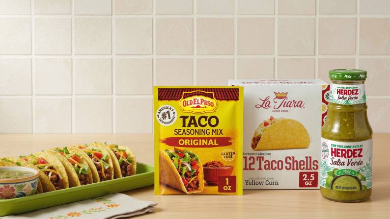 Tacos and guacamole next to a box of La Tiara tacos, a bottle of Herdez green salsa, and a packet of Old El Paso taco seasoning mix.