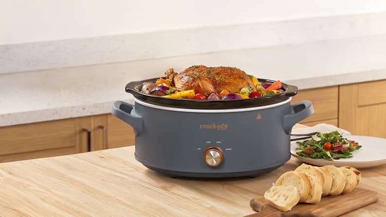 Crockpot slow cooker with roasted chicken and vegetables on a kitchen counter, with sliced bread and salad nearby.