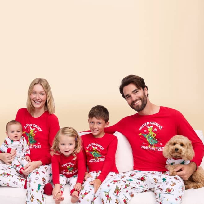 Family in matching red Mister Grinch Christmas pajamas sits on a couch with a small dog.