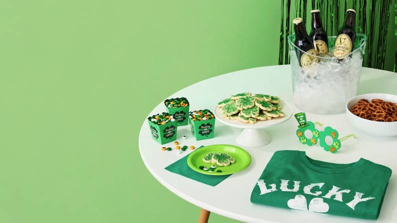 Saint Patrick’s Day snacks and accessories on table: candy, cookies, pretzels, beverages, novelty shamrock glasses, and green Lucky t-shirt.