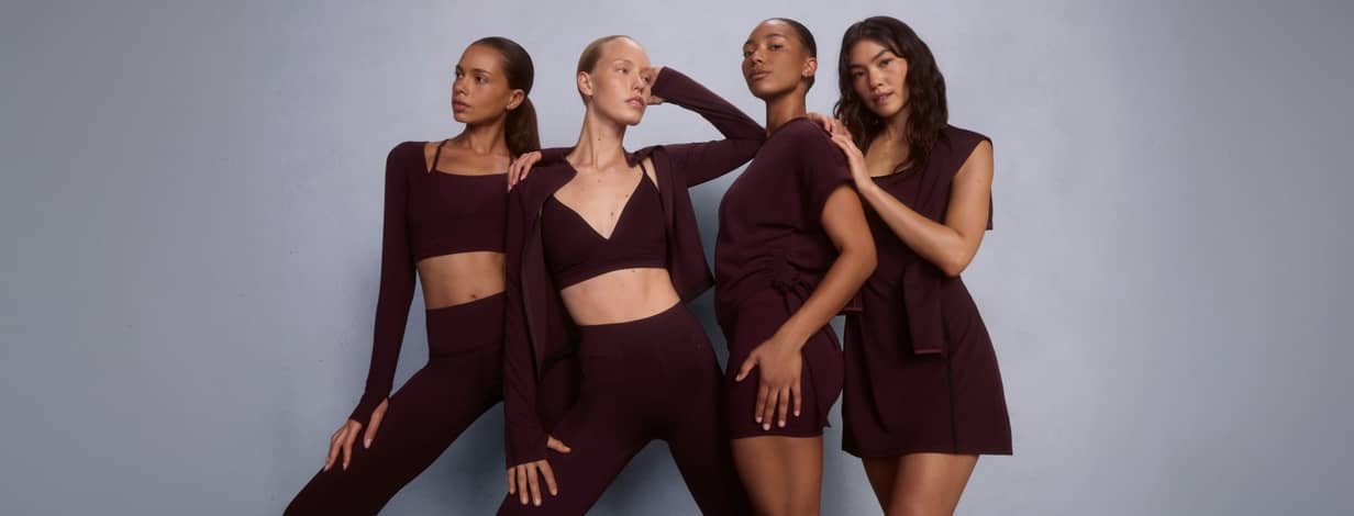 Four women posing in matching dark burgundy activewear including sports bras, leggings, tops and skirts.