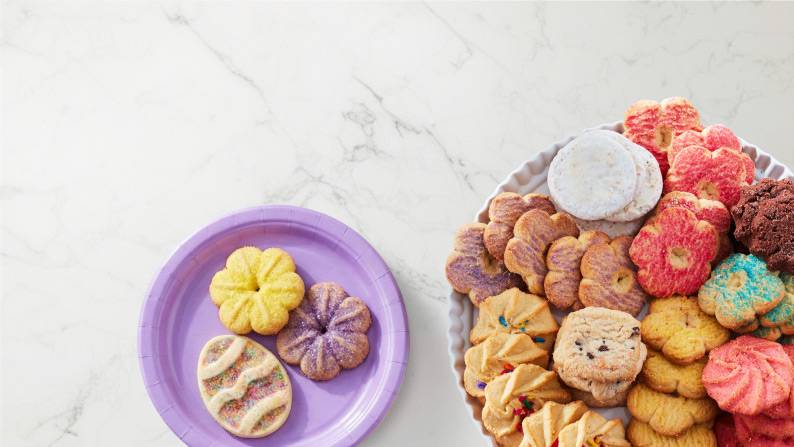 A paper plate with sugar cookies shaped like an Easter egg and flowers next to a platter of various types of cookies.