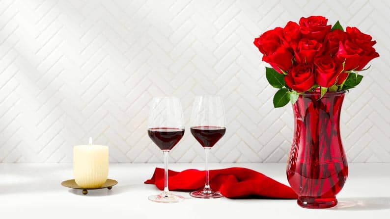 A bouquet of red roses is shown with 2 wine glasses and a candle.