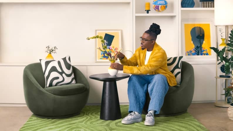 Temi Coker is seated arranging florals in a living room—two circular chairs, rug and side table are at the center. Colorful artwork is on the walls.