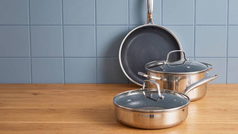 A stainless steel frying pan, pot and sauté pan with glass lids on a wood counter against blue tile. Reads: Non-toxic ceramic cookware.