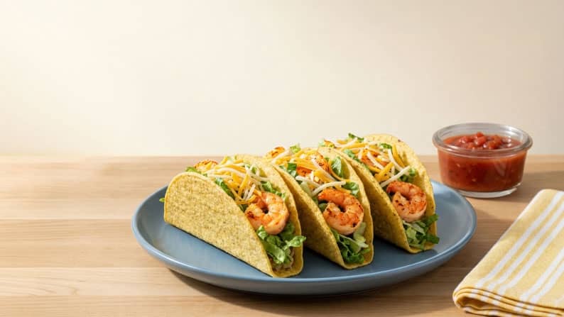 Three crispy taco shells filled with shrimp, lettuce, and cheese on a plate next to a cloth napkin and a small bowl of salsa.