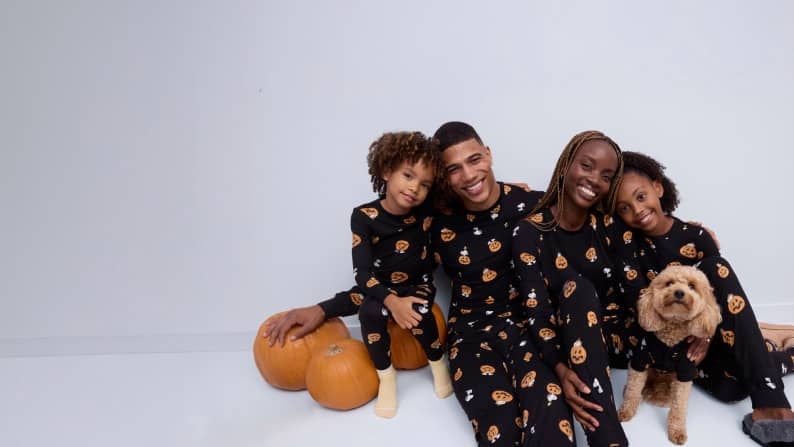 A family of four and their dog are sitting on the floor wearing matching pumpkin pajamas.