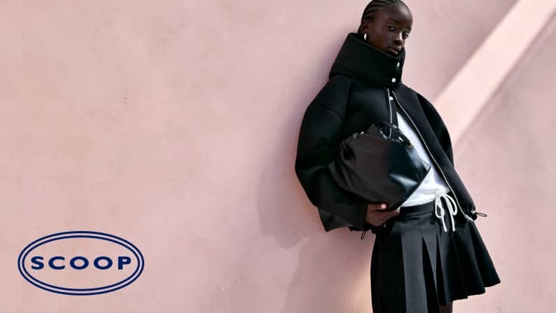 A fashion model wears a black high collar jacket and pleated skirt, holding a black bag against a soft pink wall.