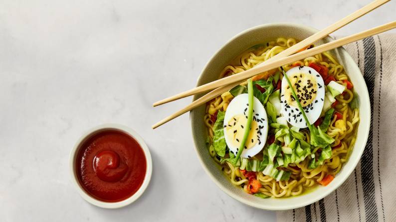 Chopsticks and a bowl of ramen topped with bok choy, hard-boiled eggs, black sesame seeds, and carrots next to a small bowl of red sauce.