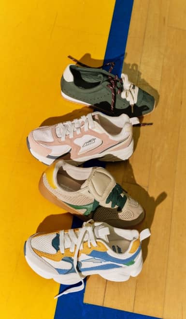 Four kids’ retro sneakers in green suede, pink mesh, khaki, and blue colorblock are lined top-to-bottom on a yellow and blue gym floor.