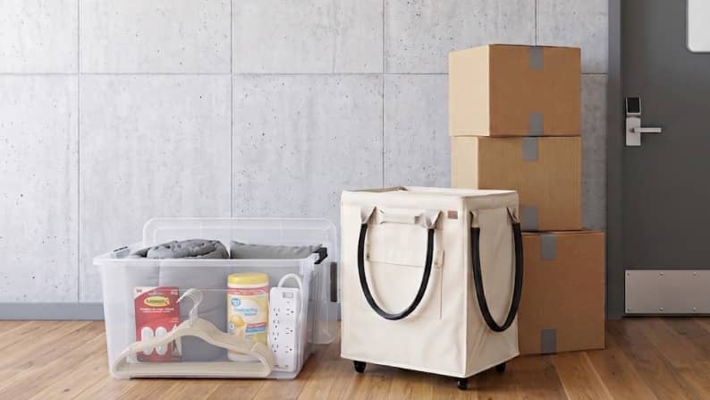 A clear plastic bin with bedding and a power strip sits next to a beige rolling hamper and a stack of cardboard moving boxes on a wood floor.