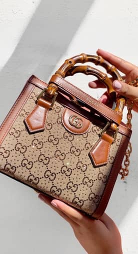 A woman is holding a small Gucci top handle bag in front of a white wall.