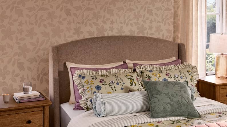 A bed with floral linens and various textured pillows sits between two wooden nightstands in a room with botanical wallpaper.