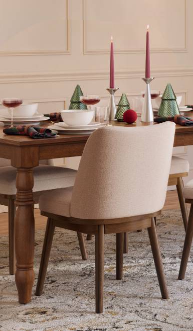 Holiday dining table with red candles, green tree decorations and place settings beside a light upholstered chair on a patterned rug.
