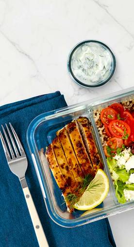 Grilled chicken, brown rice and fresh veggies are displayed in a glass storage container. A cup of yogurt dipping sauce is also shown.