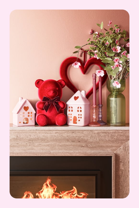 A mantel is decorated with a red plush bear, little pink houses, pink tapered candles, a flower bouquet and a red heart wall art decoration.