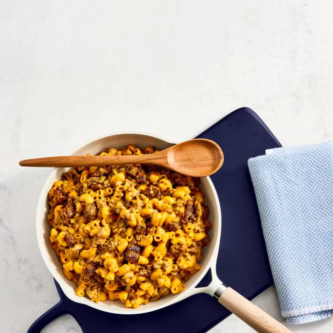 A skillet of cheesy macaroni and ground beef with a wooden spoon on a blue cutting board next to a light blue kitchen towel.
