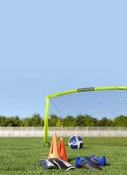 Orange cones, cleats, and shin guards sit in front of a soccer net with a ball in it.