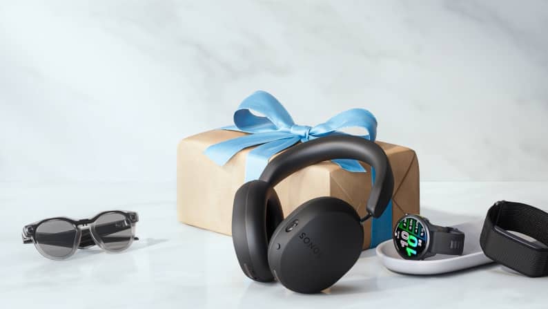 Sonos headphones, pair of smart glasses and two wrist watches on ceramic tray in front of wrapped gift box.