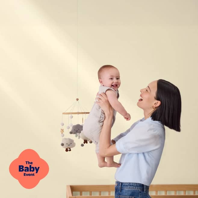Mom holding her baby smiling in front of a crib. The Baby Event badge in pink.