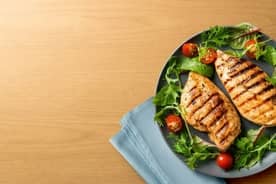 Two grilled chicken breasts on a plate with fresh greens and halved cherry tomatoes on a wooden surface next to a blue cloth napkin.