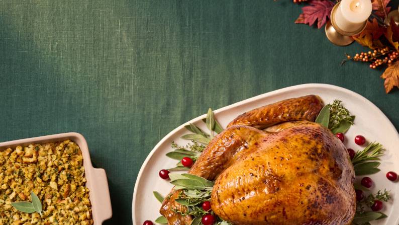 Roast turkey on a platter with herbs and cranberries beside a dish of stuffing on a green table with autumn leaves and a candle.