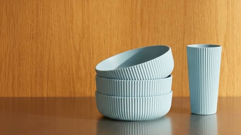 A stack of three light blue ribbed bowls next to a matching cup on a dark table. Reads: From fifty cents. Durable dinnerware.
