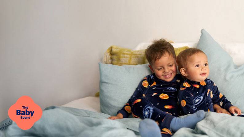 The Baby Event logo. Two little boys wearing blue planet-printed pajamas are hugging, while sitting on a bed.