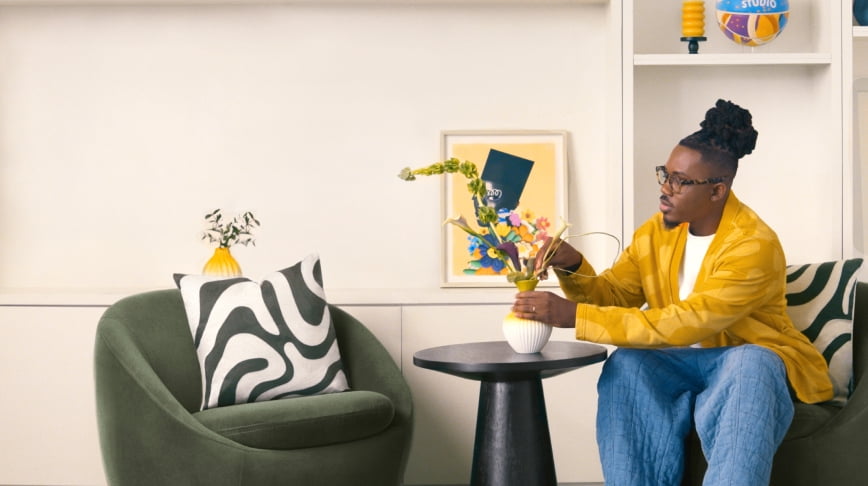 Temi Coker is seated arranging florals in a living room—two circular chairs, rug and side table are at the center. Colorful artwork is on the walls.