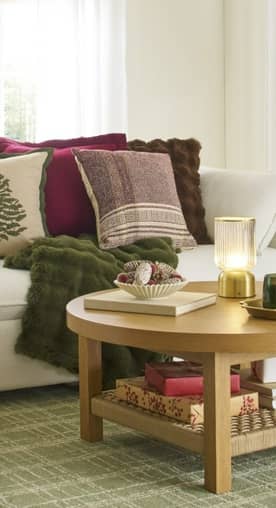 A cozy living room with a round wood coffee table and sectional sofa in the background. Throw pillows, a candle and gifts make it feel festive.