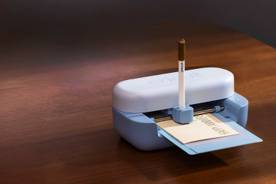 Cricut cutting machine on a table, feeding material through while a pen attachment writes and a potted plant nearby.