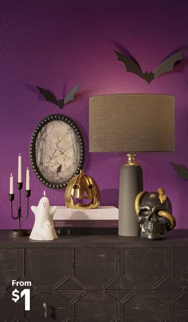 Image of a console against a purple wall with candles, ghosts, pumpkin and skull decor. Enchant all who enter. Indoor Halloween decor. Shop now.