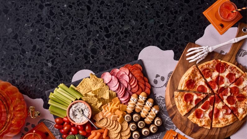 A counter full of cut vegetables, sliced meat and cheese, crackers, dip, a sliced pepperoni pizza, and orange drinks on a ghost table runner.