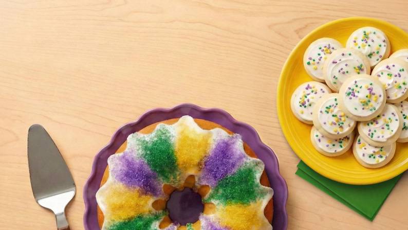 A purple, green & yellow bundt cake is shown with a plate of frosted sugar cookies.