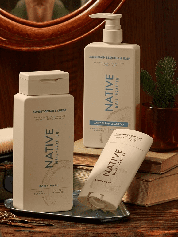 Image shows three Native personal care products—a body wash, a shampoo and a deodorant—placed on a wooden surface with a light wooden background.